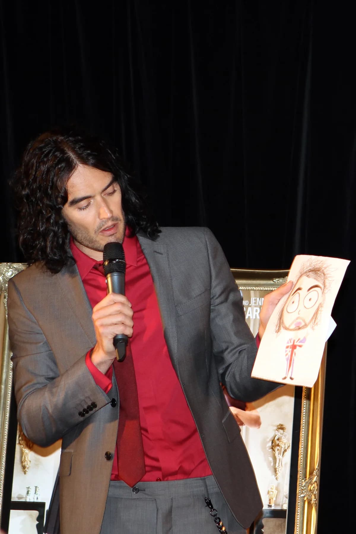 Russell Brand speaking into a microphone while holding a drawing.
